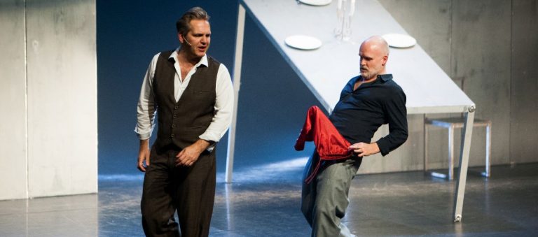 Twee acteurs zijn bezig met een expressieve scène op een podium, met een tafel en borden op de achtergrond.