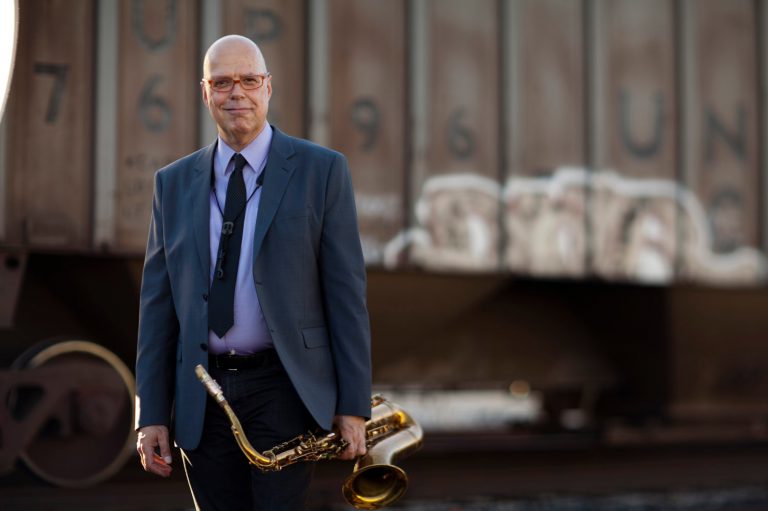 Een man in een pak houdt een saxofoon vast terwijl hij buiten poseert met een onscherpe achtergrond.