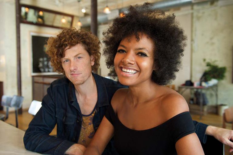 Een man en een vrouw zitten samen aan een tafel en lachen naar de camera in een gezellige omgeving.