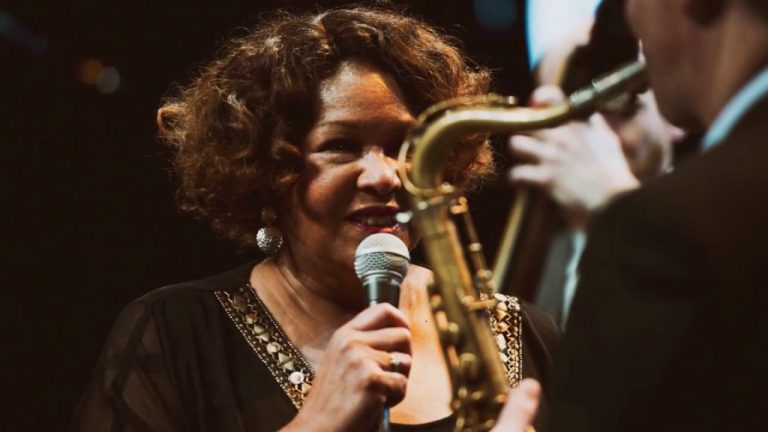 Een zangeres met krullend haar houdt een microfoon vast, terwijl een saxofonist in een optreden dichtbij haar staat.