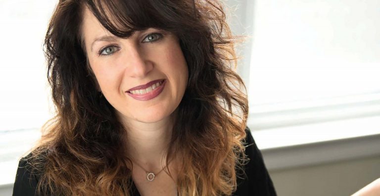 Een vrouw met krullend haar en een glimlach staat voor een lichte achtergrond.