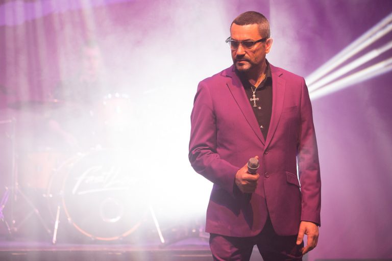 Een man in een paarse suit staat op het podium met een microfoon, omringd door kleurrijke verlichting en rook.