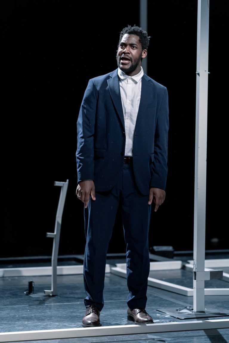 Een man in een donkerblauw kostuum staat op een podium met zaklamp achter zich.