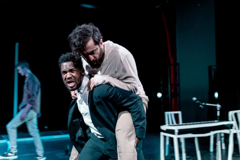 Twee acteurs voeren een intense scène op het podium uit, waarbij één acteur op de rug van de ander is gehesen.