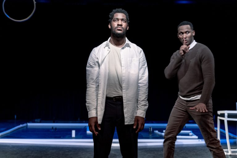 Twee mannen staan op een theaterpodium, één kijkt serieus omhoog terwijl de andere stil is en gefocust lijkt.