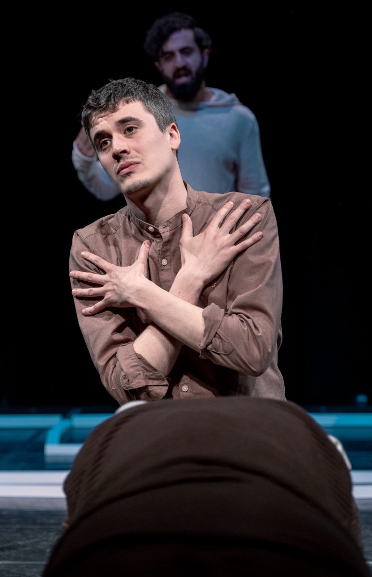 Een acteur zit op een podium met een intensieve blik, terwijl hij zijn armen om zichzelf heen slaat.