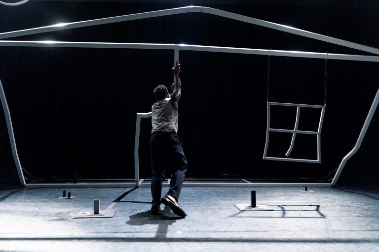 Een persoon hangt aan een structuur die de vorm van een huis nabootst in een donkere omgeving.