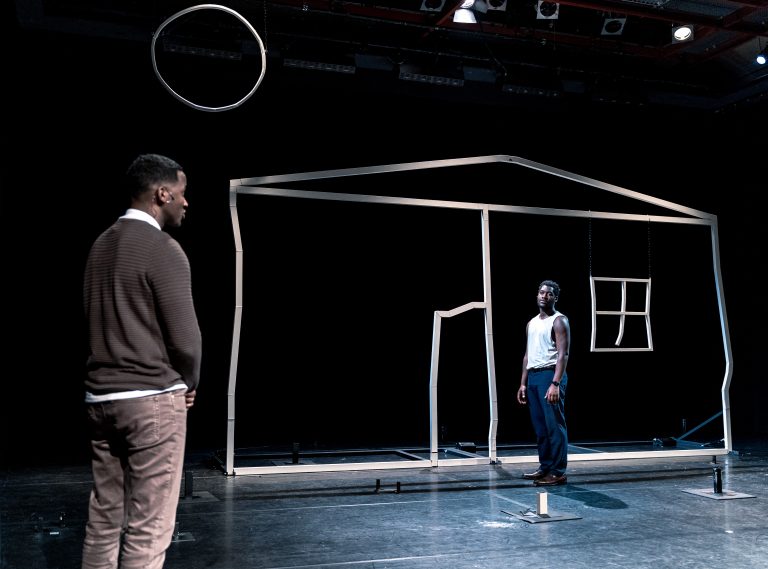 Twee acteurs staan in een donkere ruimte met een geometrische, lichtgekleurde huisconstructie op de achtergrond.