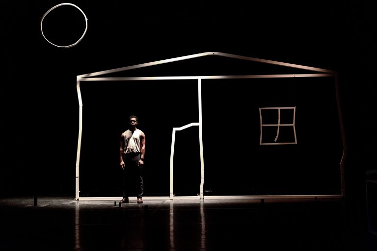Een performer staat in een donkere ruimte voor een abstracte huisstructuur gemaakt van lichtlijnen.