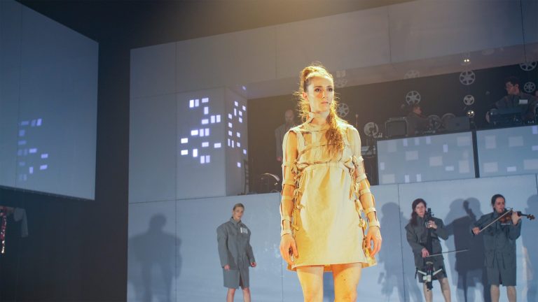 Model op een modeshow draagt een glanzende, gouden jurk, met een minimalistische achtergrond en andere modellen op de catwalk.