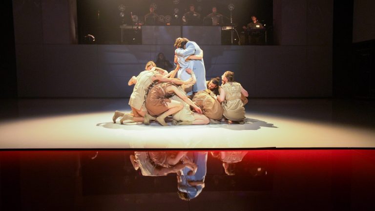 Dansers in kostuums omarmen elkaar op een podium, met een reflectie zichtbaar op de vloer.