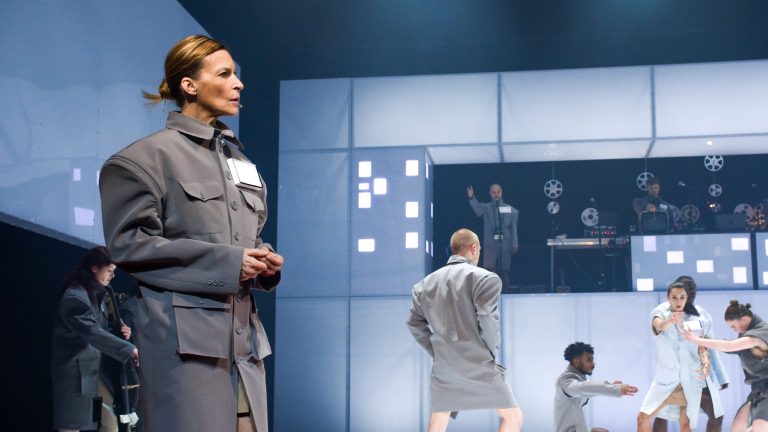 Een actrice in een grijze outfit staat op een podium met andere performers in een moderne, abstracte setting.