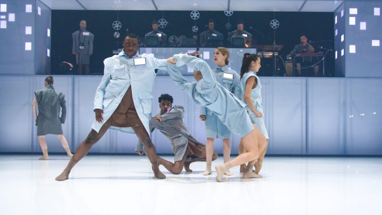 Vijf dansers voeren een choreografie uit op een ijsvloer, gekleed in lichtblauwe kostuums.