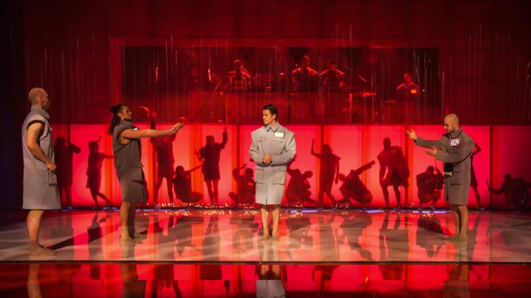 Een theatrale scène met drie performers op een podiumpartij, tegen een rode achtergrond met silhouetten van andere dansers.