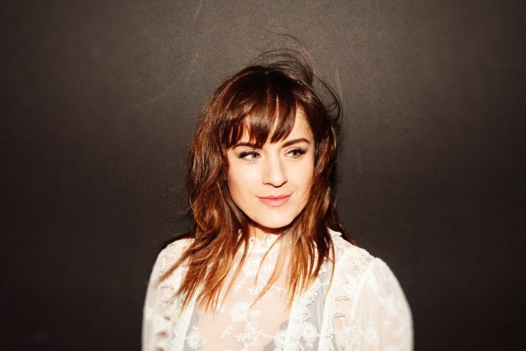 Portret van een vrouw met schouderlang bruin haar en een lichte blouse, tegen een donkere achtergrond.