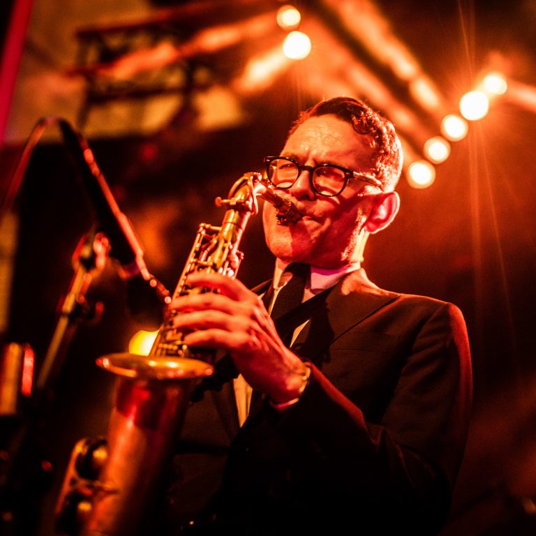 Een muzikant speelt een trompet tijdens een optreden, omgeven door warme sfeerverlichting.