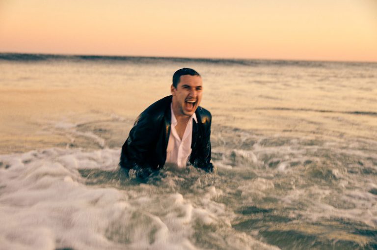 Een man in een zwart jasje die met een mond vol water in de branding staat en schreeuwt tijdens zonsondergang.