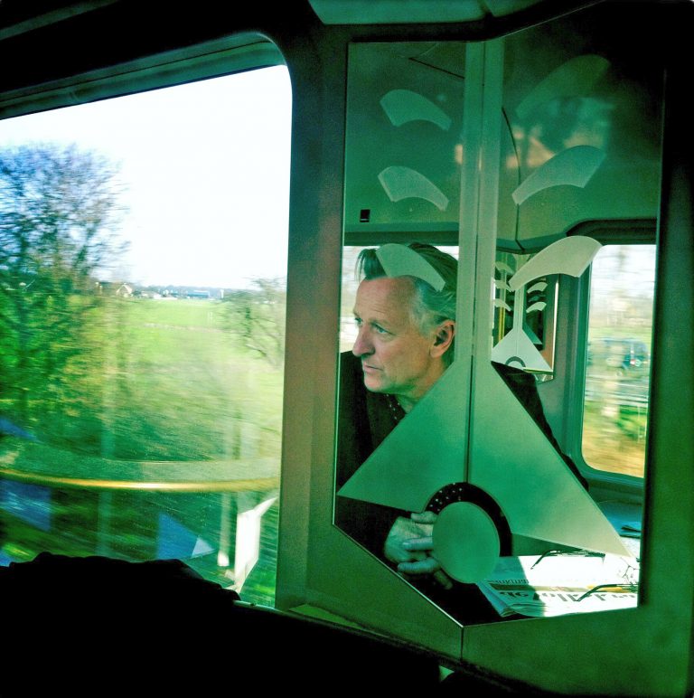 Een man kijkt met een ontspannen uitdrukking naar buiten door een treinenraam, omringd door groen landschap.