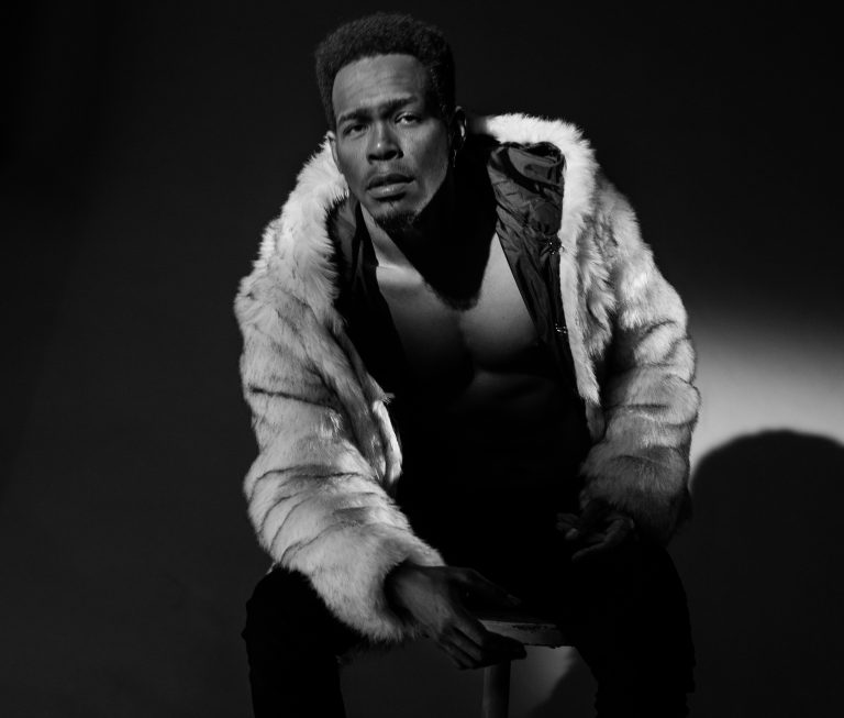 Een man in een stijlvol bontjack zit op een stoel tegen een donkere achtergrond.