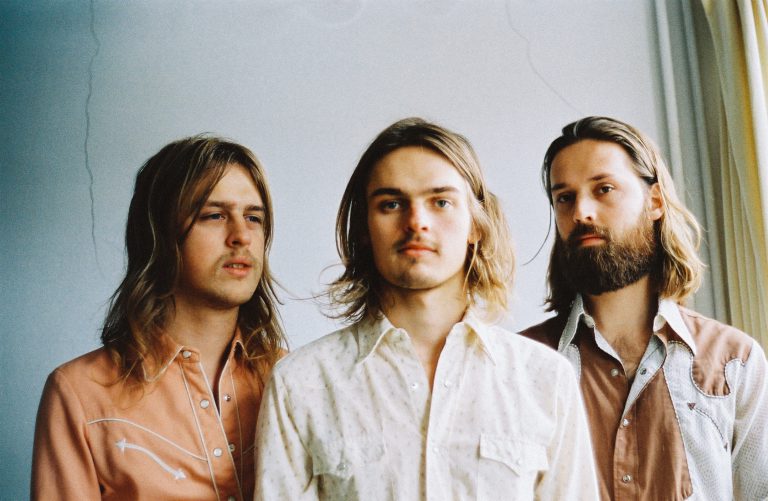 Drie mannen met lang haar staan in een lichte setting, gekleed in casual overhemden.