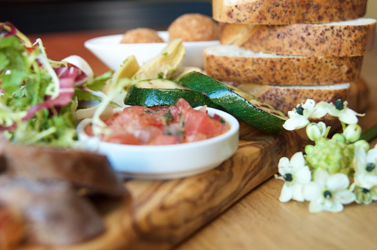Een houten plank met gesneden groenten, een schaaltje met salsa en verschillende soorten brood op de achtergrond.
