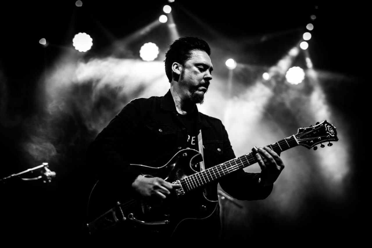 Een muzikant speelt gitaar op een podium met lichten op de achtergrond, in zwart-wit.