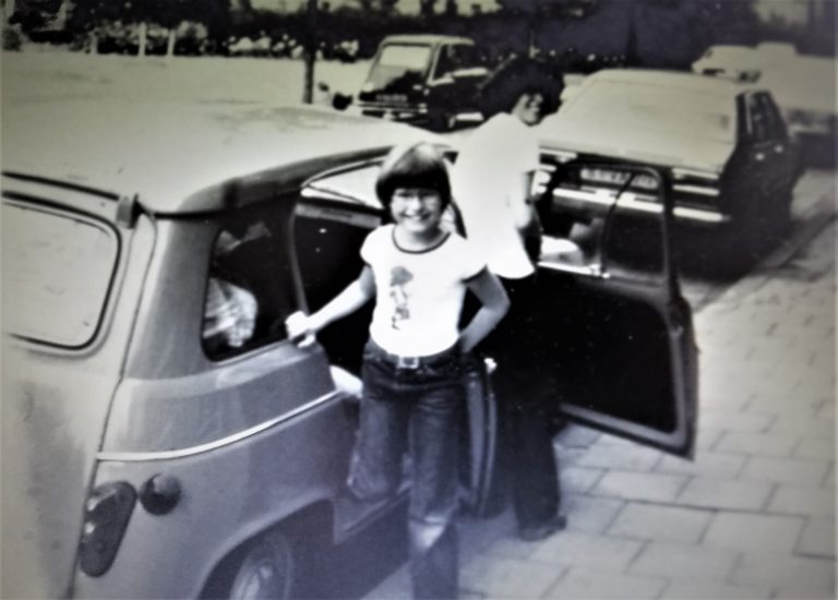 Een jonge persoon staat naast een auto en poseert, terwijl een andere persoon binnenin de auto bezig is.