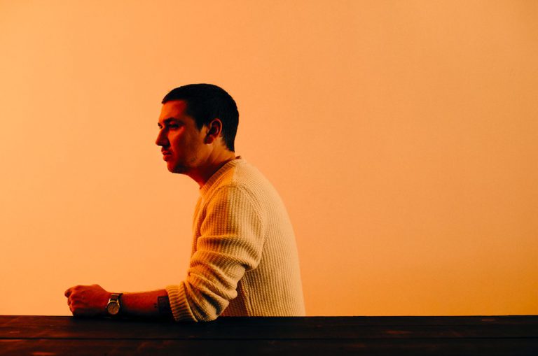 Een persoon in profiel staat aan een tafel, met een oranje achtergrond.