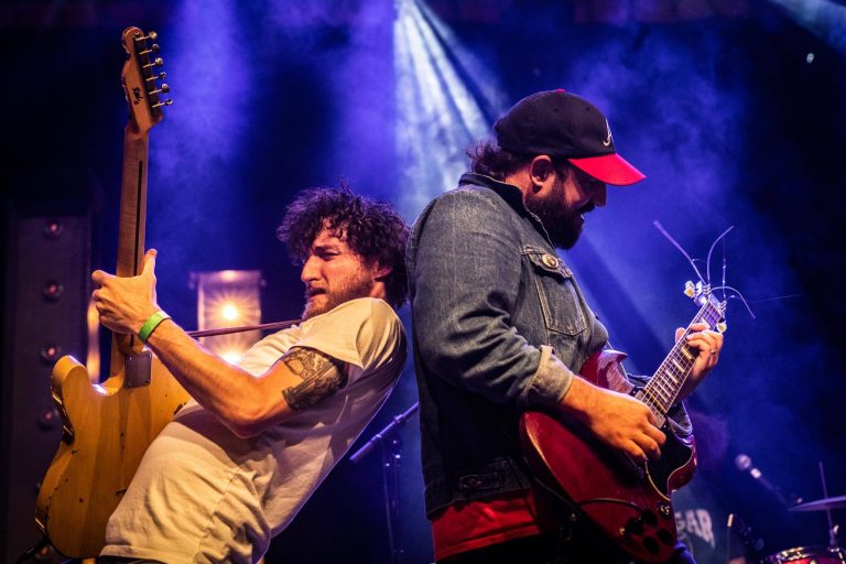 Twee mannen met hun ruggen tegen elkaar aan spelend op gitaren