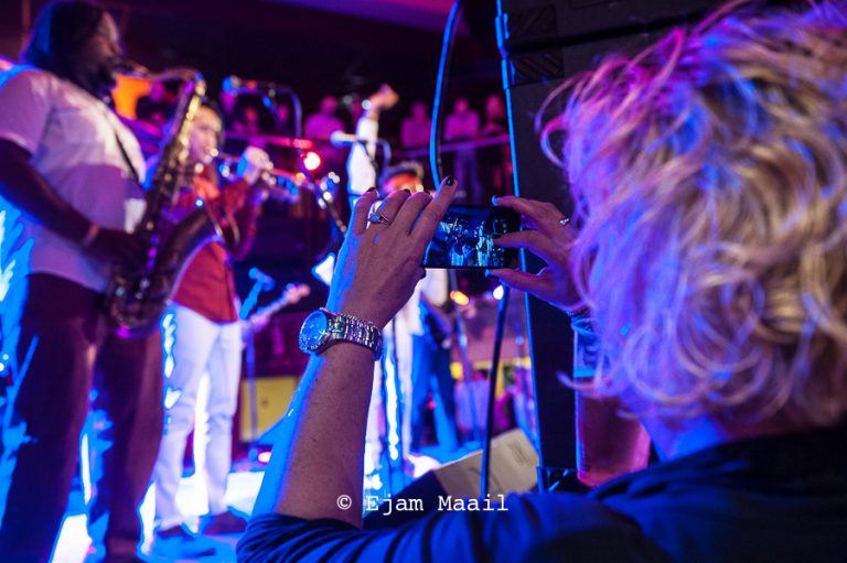 Een persoon maakt een foto van een band die optreedt in een sfeervolle, kleurrijke omgeving.