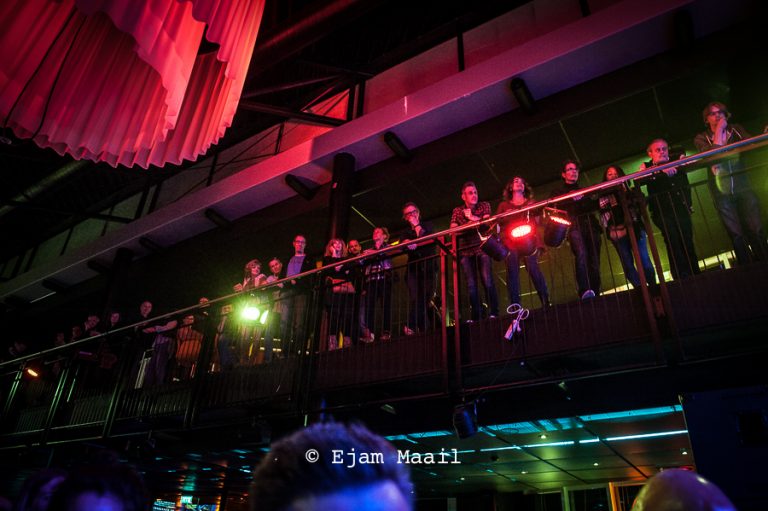 Een groep mensen staat op een balkon in een levendige, met lichtjes verlichte ruimte.