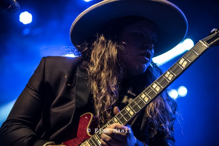 Een muzikant met lang haar speelt gitaar op een podium in een blauwe lichtomgeving.