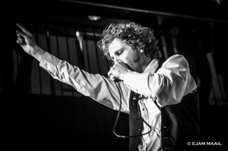 Een muzikant zingt met één arm omhoog, terwijl hij in een zwart-wit setting op het podium staat.