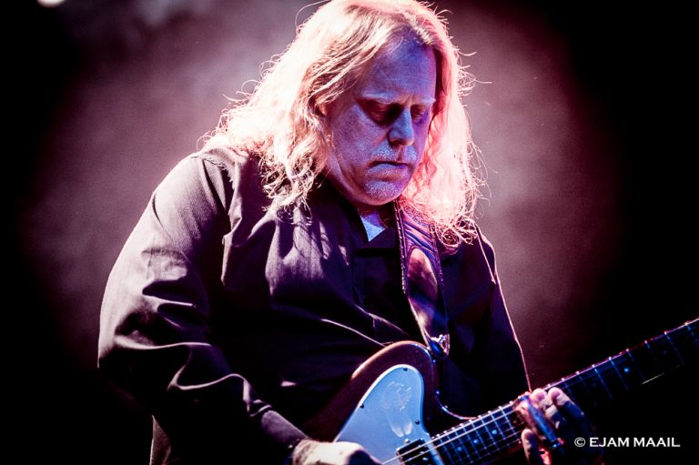 Een muzikant speelt op een elektrische gitaar met een intense uitstraling tegen een sfeervolle, verlichte achtergrond.