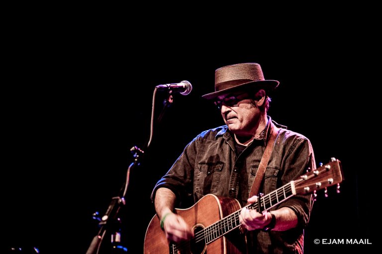Een man met een gitaar en hoed die een optreden geeft op een podium met dim licht.
