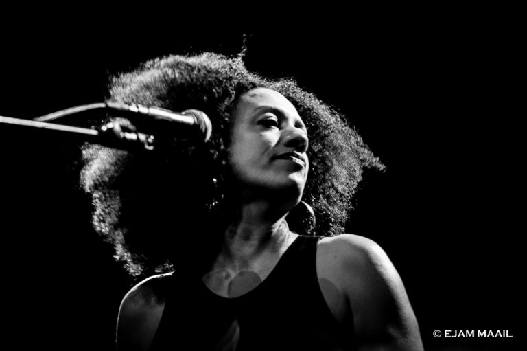 Een vrouw met een volle afro en microphone, kijkt contemplatief omhoog in een zwart-wit setting.