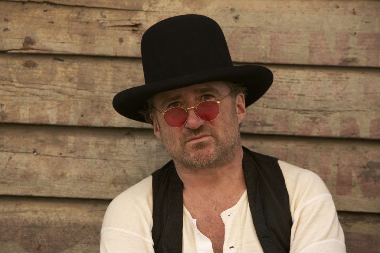 Een man met een hoge hoed en roze zonnebril zit tegen een houten achtergrond.