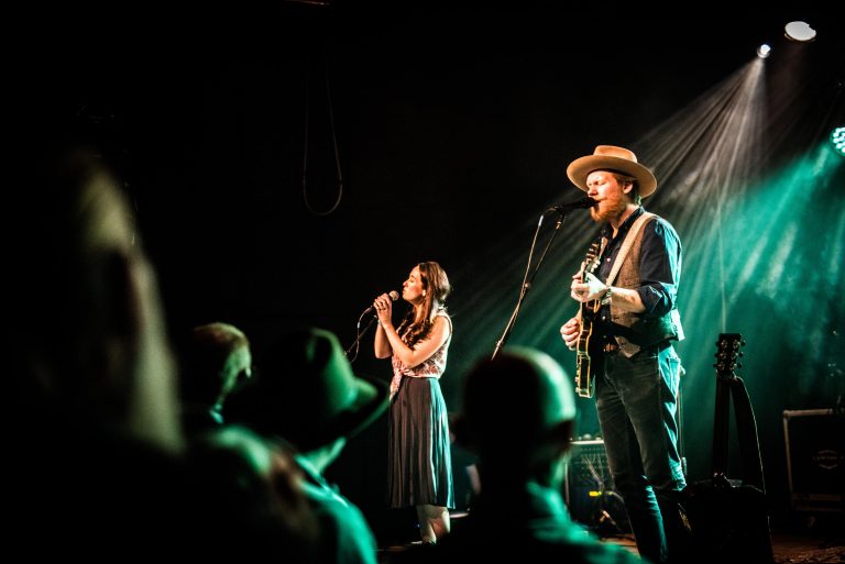 Twee muzikanten performen op een podium met groene belichting terwijl het publiek naar hen kijkt.
