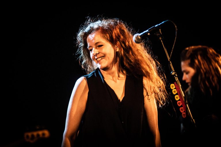 Zangeres met krullend haar, glimlachend voor een microfoon op het podium.