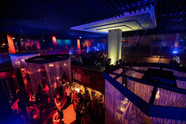 Een levendige nachtclub met felgekleurde verlichting en een drukte van mensen op de dansvloer en rond de bar.