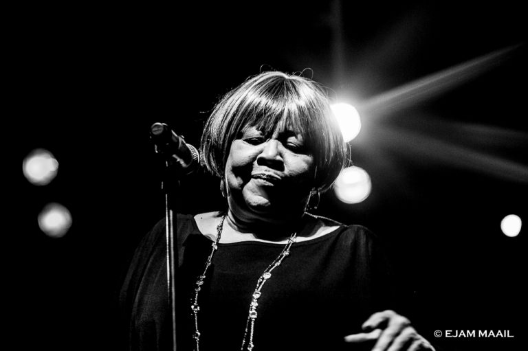 Een vrouw zingt met een microfoon in zwart-wit, terwijl lichteffecten op de achtergrond zichtbaar zijn.