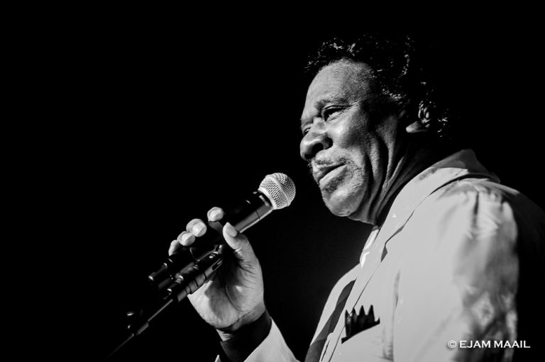 Een man in een lichtkostuum houdt een microfoon vast terwijl hij glimlacht tijdens een optreden.