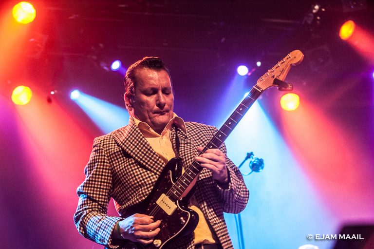 Een muzikant in een gestreepte jas speelt op een zwarte gitaar onder kleurrijke podiumverlichting.