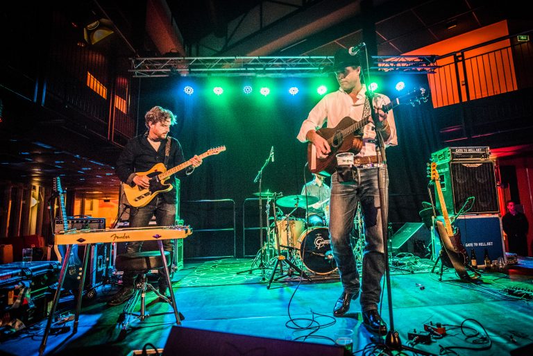 Een band performt op het podium met groene verlichting, terwijl een gitarist en een zanger met gitaar zichtbaar zijn.