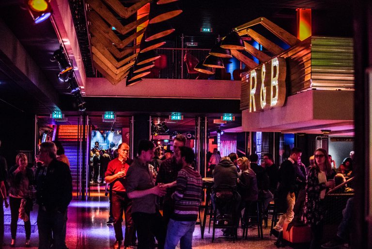 Een drukke bar met kleurrijke verlichting en mensen die gezellig met elkaar praten.