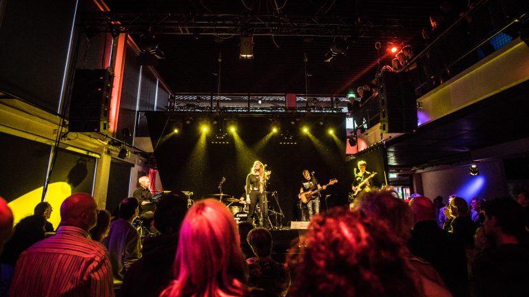 Een muziekband treedt op op een podium met heldere lichteffecten, terwijl het publiek toekijkt.