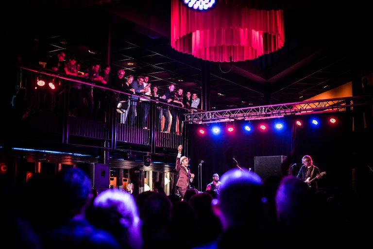 Een muzikant staat op het podium van een levendige concertzaal, omringd door licht en een enthousiaste menigte.
