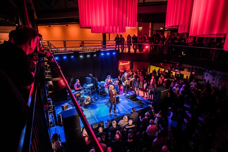 Een overzicht van een live muziekoptreden in een sfeervolle locatie met een druk publiek en kleurrijke verlichting.