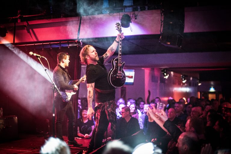 Een muzikant staat op het podium met een gitaar in de lucht, terwijl het publiek enthousiast naar hem kijkt.