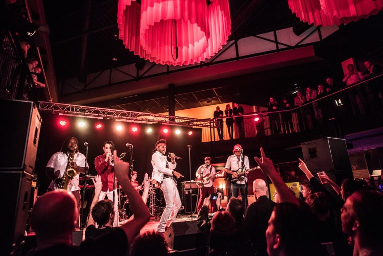 Een energieke liveband speelt op een podium, omringd door een enthousiaste menigte en sfeervolle rode verlichting.